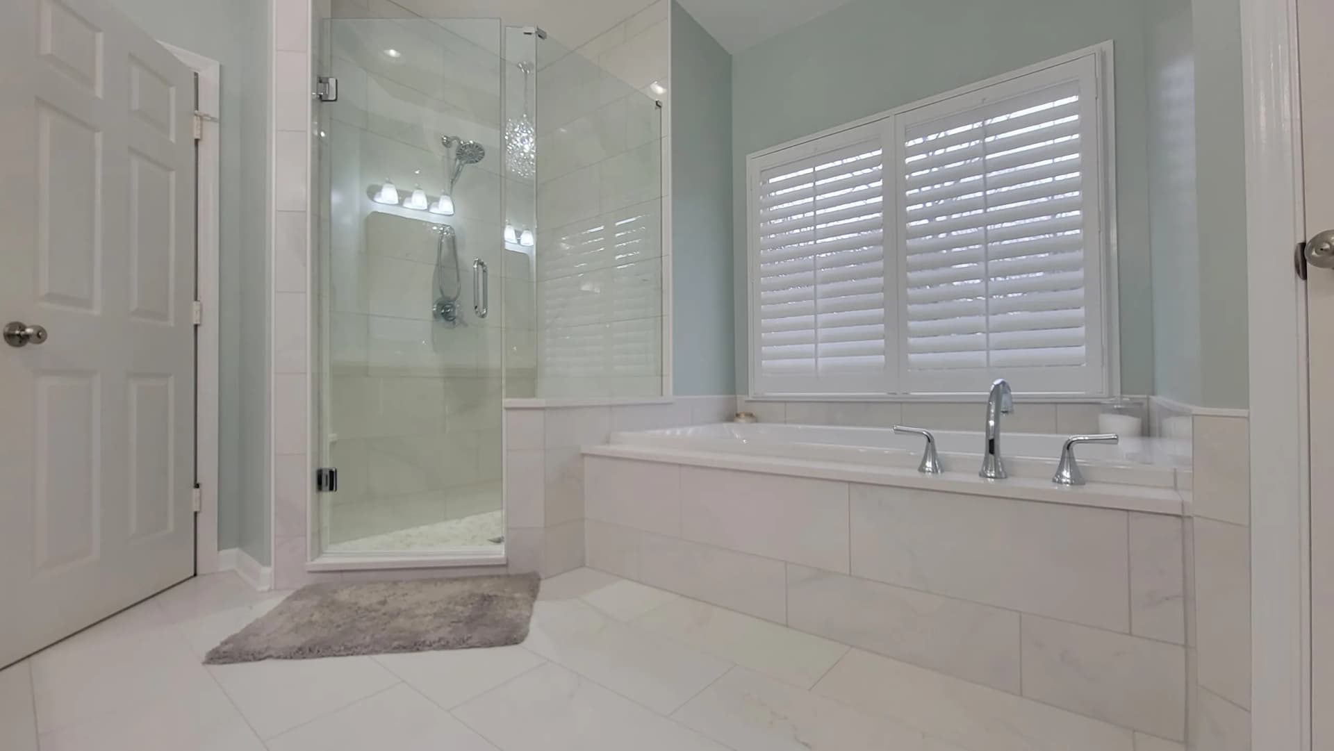 Bright bathroom with tiled shower, built-in tub, large window with shutters, and white tile flooring